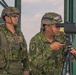Two military personnel in camouflage uniforms are positioned at what appears to be an observation post or elevated platform. One soldier is looking through a spotting scope mounted on a tripod, while the other stands watch. Both wear tactical helmets with goggles and display unit patches on their uniforms.