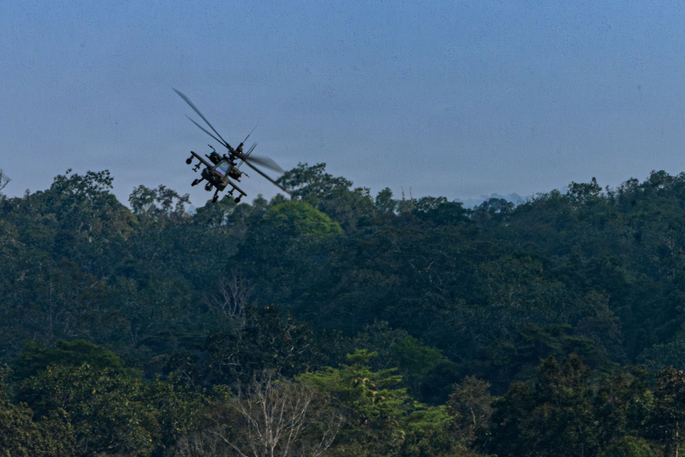 A military attack helicopter is flying low over a dense tropical forest canopy. The helicopter appears to be an AH-64 Apache or similar attack variant, identifiable by its tandem cockpit configuration and weapons pylons on the stub wings. The image was taken from a distance, with the helicopter silhouetted against a hazy blue sky.