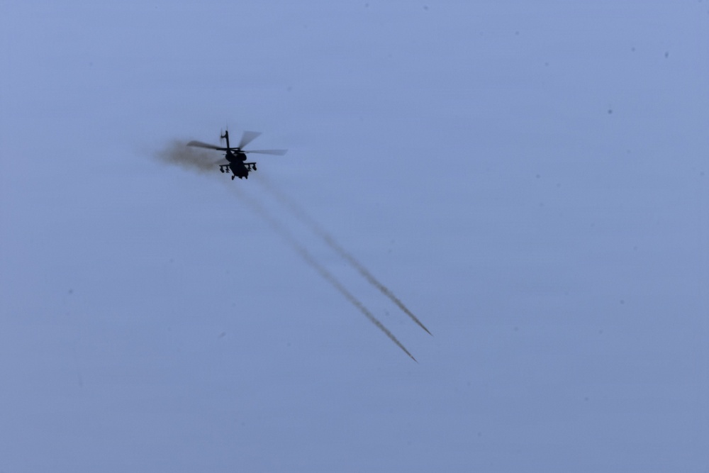 A military attack helicopter is captured in silhouette against an overcast, pale blue-grey sky. The helicopter has just fired what appear to be two rockets or missiles, visible as twin smoke trails extending diagonally downward from the aircraft. The image is taken from a significant distance, rendering the helicopter as a dark silhouette.