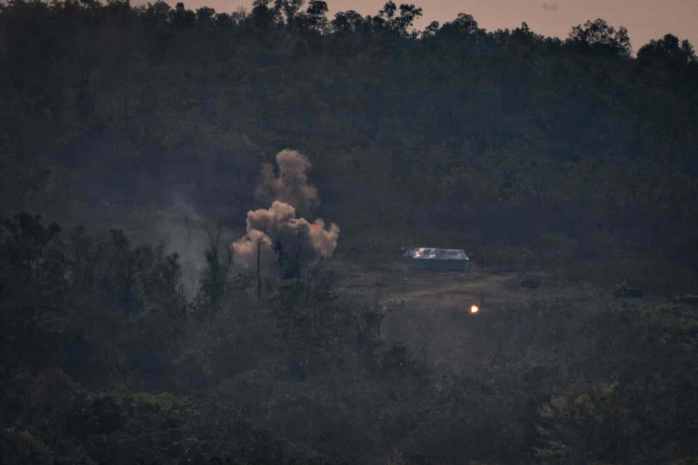 A distant telephoto image captured at dusk or dawn showing an explosion or detonation occurring near a small structure in a forested hillside area. A large plume of smoke and debris is visible rising from the blast site, with a separate bright light source visible to the right of the building. The scene suggests an active incident involving fire, explosion, or demolition in a rural or semi-rural environment.