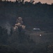 A distant telephoto image captured at dusk or dawn showing an explosion or detonation occurring near a small structure in a forested hillside area. A large plume of smoke and debris is visible rising from the blast site, with a separate bright light source visible to the right of the building. The scene suggests an active incident involving fire, explosion, or demolition in a rural or semi-rural environment.
