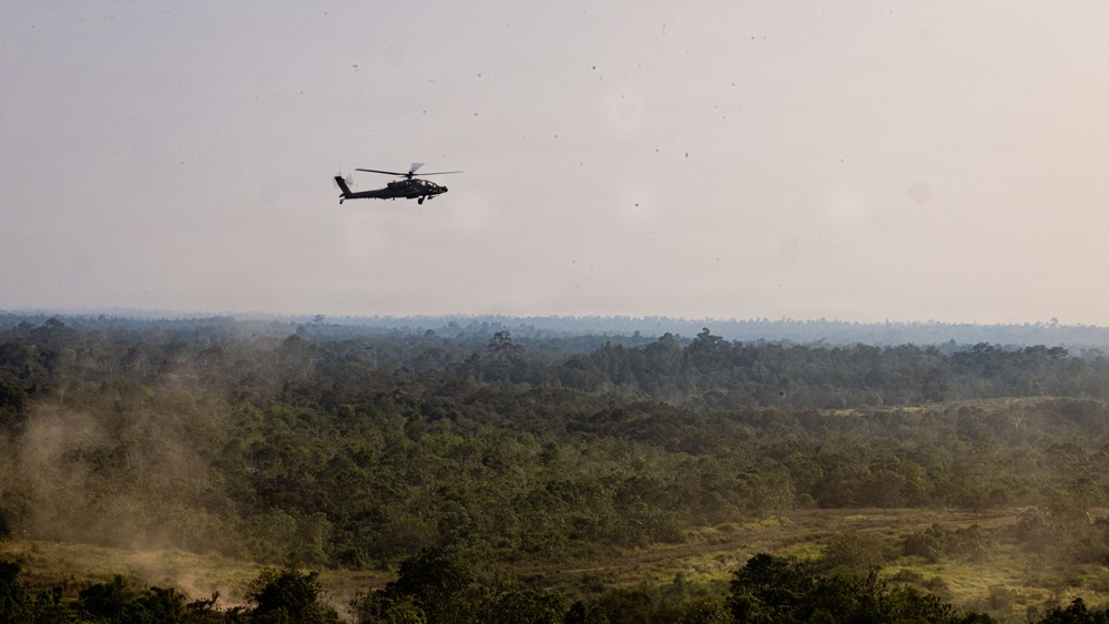 A military attack helicopter, consistent with an AH-64 Apache, flies low over a dense tropical forest landscape. The terrain below features a mix of jungle canopy and cleared areas with visible dust or smoke. The hazy atmospheric conditions suggest either dry season conditions or nearby burning activity.