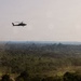 A military attack helicopter, consistent with an AH-64 Apache, flies low over a dense tropical forest landscape. The terrain below features a mix of jungle canopy and cleared areas with visible dust or smoke. The hazy atmospheric conditions suggest either dry season conditions or nearby burning activity.