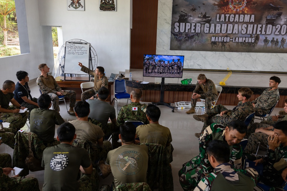 A multinational military briefing or training session is taking place at the 'Super Garuda Shield' exercise at MARFOR-Lampung, Indonesia. Personnel from multiple nations including the United States, Japan, and Indonesia are gathered in a room while a presenter points to a whiteboard. A television screen displays soldiers in the field, likely as part of the instructional content.
