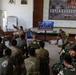 A multinational military briefing or training session is taking place at the 'Super Garuda Shield' exercise at MARFOR-Lampung, Indonesia. Personnel from multiple nations including the United States, Japan, and Indonesia are gathered in a room while a presenter points to a whiteboard. A television screen displays soldiers in the field, likely as part of the instructional content.
