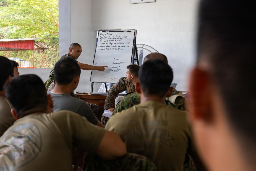 A military instructor is conducting a briefing or training session for a group of soldiers/marines in what appears to be an outdoor or semi-open classroom setting. The instructor is pointing at a whiteboard/flip chart labeled 'Focus Points' that contains written bullet points and a hand-drawn diagram. The attendees are seated and wearing various military uniforms, suggesting a multinational or joint training exercise.