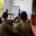 A military instructor is conducting a briefing or training session for a group of soldiers/marines in what appears to be an outdoor or semi-open classroom setting. The instructor is pointing at a whiteboard/flip chart labeled 'Focus Points' that contains written bullet points and a hand-drawn diagram. The attendees are seated and wearing various military uniforms, suggesting a multinational or joint training exercise.