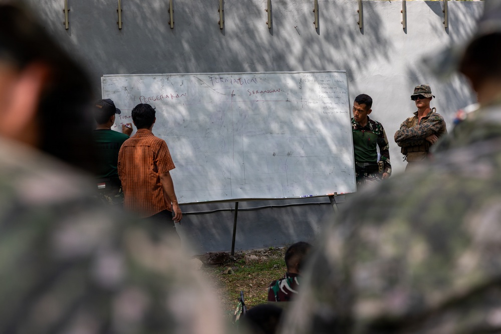 A military training or briefing session is taking place outdoors, with personnel gathered around a whiteboard. The whiteboard contains text in what appears to be Indonesian, including the words 'PERGERAKAN' (movement), 'SERANGAN' (attack), and 'FORMATION'. At least two uniformed military personnel — one in camouflage fatigues and one in what appears to be a Western-style military uniform with a cap — are observing participants writing on the board.