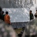 A military training or briefing session is taking place outdoors, with personnel gathered around a whiteboard. The whiteboard contains text in what appears to be Indonesian, including the words 'PERGERAKAN' (movement), 'SERANGAN' (attack), and 'FORMATION'. At least two uniformed military personnel — one in camouflage fatigues and one in what appears to be a Western-style military uniform with a cap — are observing participants writing on the board.