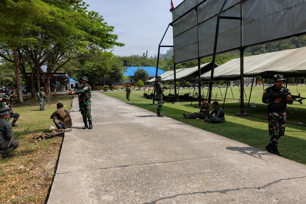 The image shows military personnel in camouflage uniforms gathered on a grassy compound, likely a military base or training facility. Several soldiers are positioned along a concrete pathway, with some standing and others seated on the grass. Large canvas shade structures are erected on the right side of the frame, and lush trees line the left side.