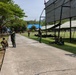The image shows military personnel in camouflage uniforms gathered on a grassy compound, likely a military base or training facility. Several soldiers are positioned along a concrete pathway, with some standing and others seated on the grass. Large canvas shade structures are erected on the right side of the frame, and lush trees line the left side.