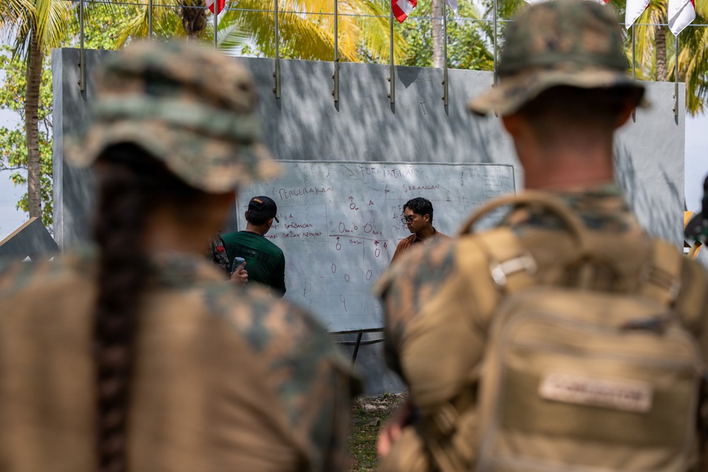 Military personnel in camouflage uniforms observe a briefing or planning session in front of a whiteboard with text and diagrams written in what appears to be Indonesian or Malay language. Two civilian-dressed individuals stand at the whiteboard, which contains tactical or organizational information. The setting appears to be an outdoor or semi-outdoor location with tropical vegetation visible in the background.