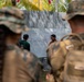 Military personnel in camouflage uniforms observe a briefing or planning session in front of a whiteboard with text and diagrams written in what appears to be Indonesian or Malay language. Two civilian-dressed individuals stand at the whiteboard, which contains tactical or organizational information. The setting appears to be an outdoor or semi-outdoor location with tropical vegetation visible in the background.