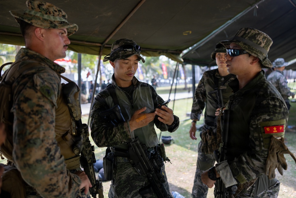 A group of military personnel from at least two different nations are gathered under a field tent canopy. One soldier is looking at a mobile phone while others observe, suggesting a coordination or communication activity. The soldiers are equipped with rifles, tactical gear, and wear distinct camouflage uniforms indicating a joint military exercise or operation.