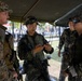 A group of military personnel from at least two different nations are gathered under a field tent canopy. One soldier is looking at a mobile phone while others observe, suggesting a coordination or communication activity. The soldiers are equipped with rifles, tactical gear, and wear distinct camouflage uniforms indicating a joint military exercise or operation.