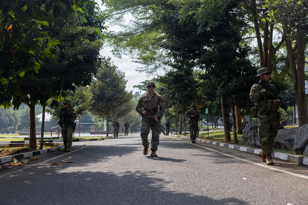A group of armed military personnel in combat gear are conducting a patrol or movement exercise along a tree-lined road. The soldiers appear to represent multiple nationalities, suggesting a joint military exercise or training operation. The setting appears to be a military base or installation in a tropical environment.