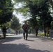 A group of armed military personnel in combat gear are conducting a patrol or movement exercise along a tree-lined road. The soldiers appear to represent multiple nationalities, suggesting a joint military exercise or training operation. The setting appears to be a military base or installation in a tropical environment.