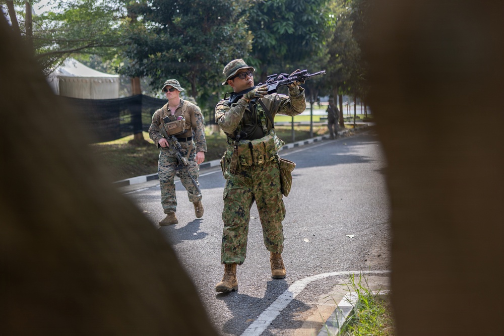 Two armed military personnel are conducting what appears to be a tactical training exercise on a paved road in a wooded or park-like setting. The foreground soldier is aiming a rifle upward while the background soldier moves in a supporting position. The setting, mixed camouflage patterns, and controlled environment suggest a joint military exercise.