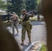 Two armed military personnel are conducting what appears to be a tactical training exercise on a paved road in a wooded or park-like setting. The foreground soldier is aiming a rifle upward while the background soldier moves in a supporting position. The setting, mixed camouflage patterns, and controlled environment suggest a joint military exercise.