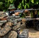 Two military personnel in different camouflage uniforms are aiming rifles in an outdoor training or exercise setting. The foreground soldier wears MARPAT (US Marine) digital camouflage, while the background soldier wears a woodland/tropical pattern consistent with Southeast Asian military forces. Both are in shooting stances with their weapons raised.