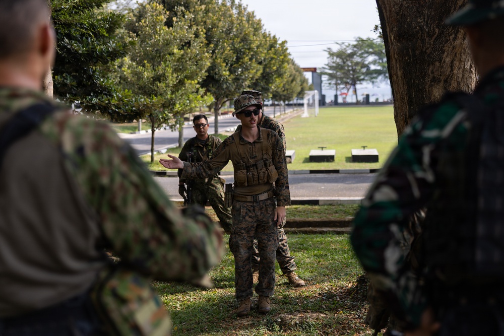 A military briefing or instruction session is taking place outdoors in a park-like setting. A central figure in US Marine Corps camouflage uniform with tactical vest and cover is addressing a small group of personnel wearing mixed camouflage patterns. The setting appears to be a military installation with manicured grounds, trees, and open fields visible in the background.