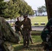 A military briefing or instruction session is taking place outdoors in a park-like setting. A central figure in US Marine Corps camouflage uniform with tactical vest and cover is addressing a small group of personnel wearing mixed camouflage patterns. The setting appears to be a military installation with manicured grounds, trees, and open fields visible in the background.