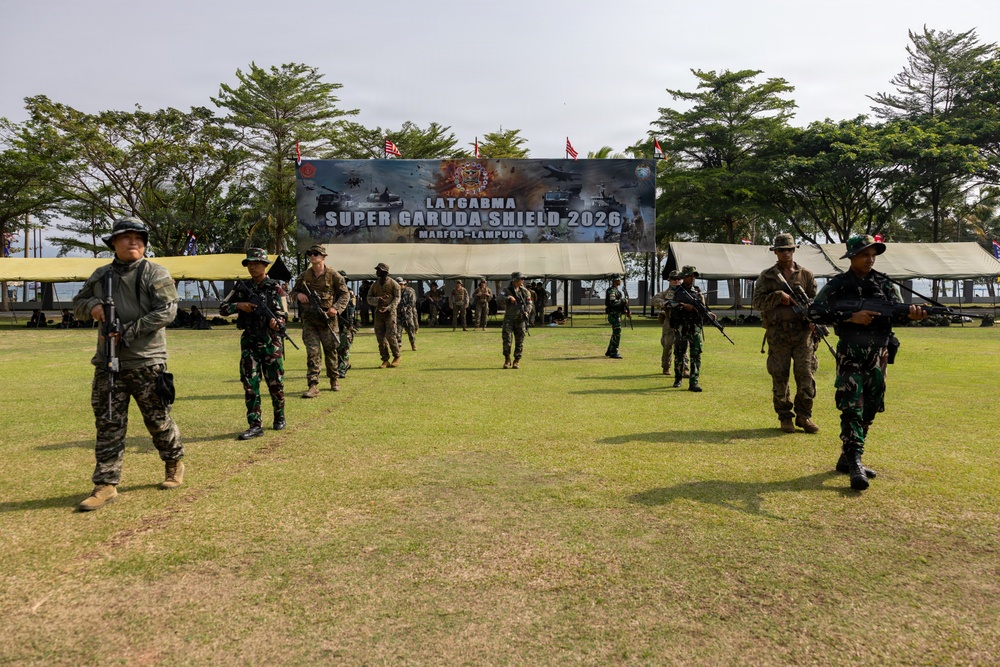 The image depicts a military exercise called 'Super Garuda Shield 2026' held at MARFOR-Lampung, Indonesia. Multiple soldiers in mixed uniforms — Indonesian TNI (green camouflage) and what appear to be U.S. Marines (tan/brown camouflage) — are conducting a joint training exercise on an open grass field. A large banner identifying the exercise is prominently displayed in the background.