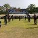 The image depicts a military exercise called 'Super Garuda Shield 2026' held at MARFOR-Lampung, Indonesia. Multiple soldiers in mixed uniforms — Indonesian TNI (green camouflage) and what appear to be U.S. Marines (tan/brown camouflage) — are conducting a joint training exercise on an open grass field. A large banner identifying the exercise is prominently displayed in the background.