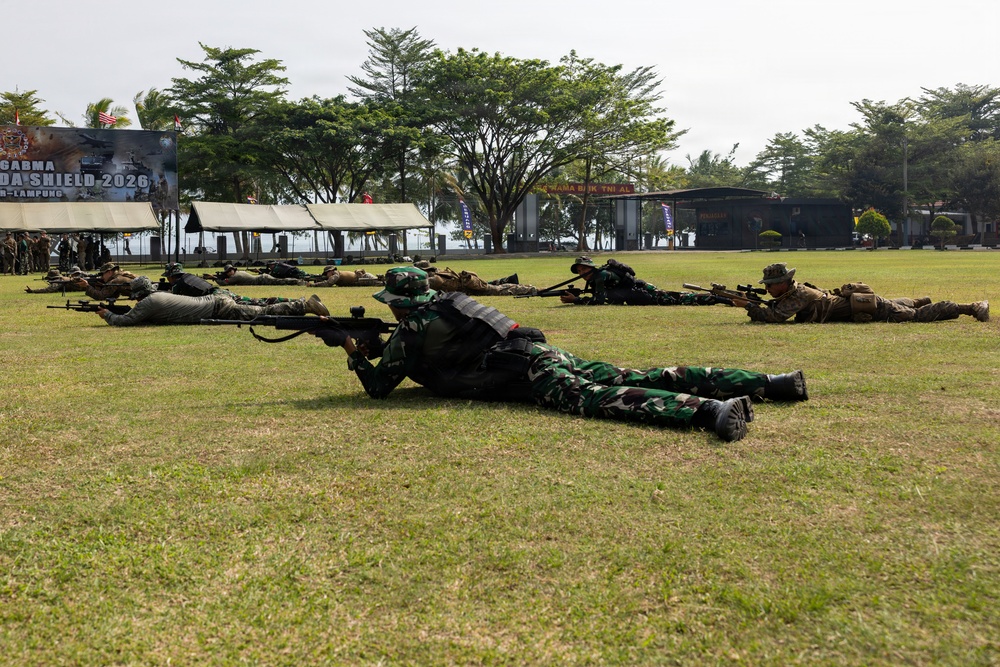 Military personnel in prone firing positions on a grass field during what appears to be a joint training exercise. Soldiers wearing two distinct uniforms — Indonesian TNI woodland camouflage and US-style tan/multicam combat gear — are lined up side by side aiming rifles downrange. A banner in the background references 'GARUDA SHIELD 2026' and 'Lampung,' indicating a bilateral or multinational military exercise.