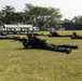 Military personnel in prone firing positions on a grass field during what appears to be a joint training exercise. Soldiers wearing two distinct uniforms — Indonesian TNI woodland camouflage and US-style tan/multicam combat gear — are lined up side by side aiming rifles downrange. A banner in the background references 'GARUDA SHIELD 2026' and 'Lampung,' indicating a bilateral or multinational military exercise.