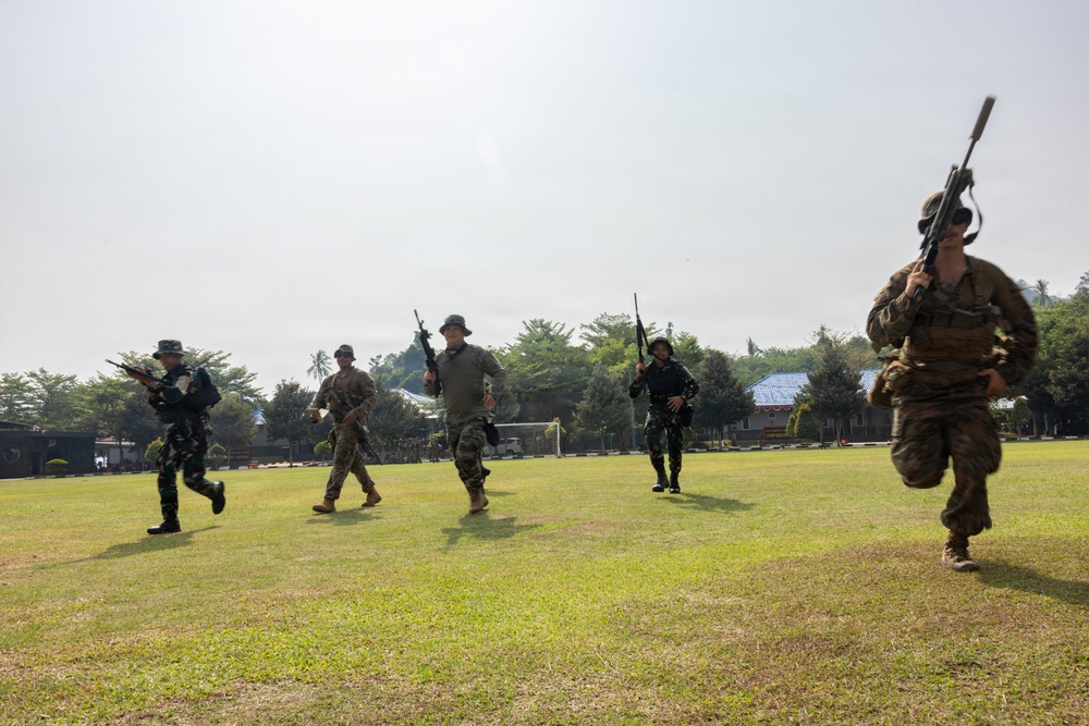 A group of five armed military personnel in mixed camouflage uniforms are conducting a tactical movement drill across an open grass field. The soldiers appear to represent at least two different national militaries, suggesting a joint training exercise. The setting appears to be a military installation or training ground with buildings and trees visible in the background.