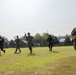 A group of five armed military personnel in mixed camouflage uniforms are conducting a tactical movement drill across an open grass field. The soldiers appear to represent at least two different national militaries, suggesting a joint training exercise. The setting appears to be a military installation or training ground with buildings and trees visible in the background.