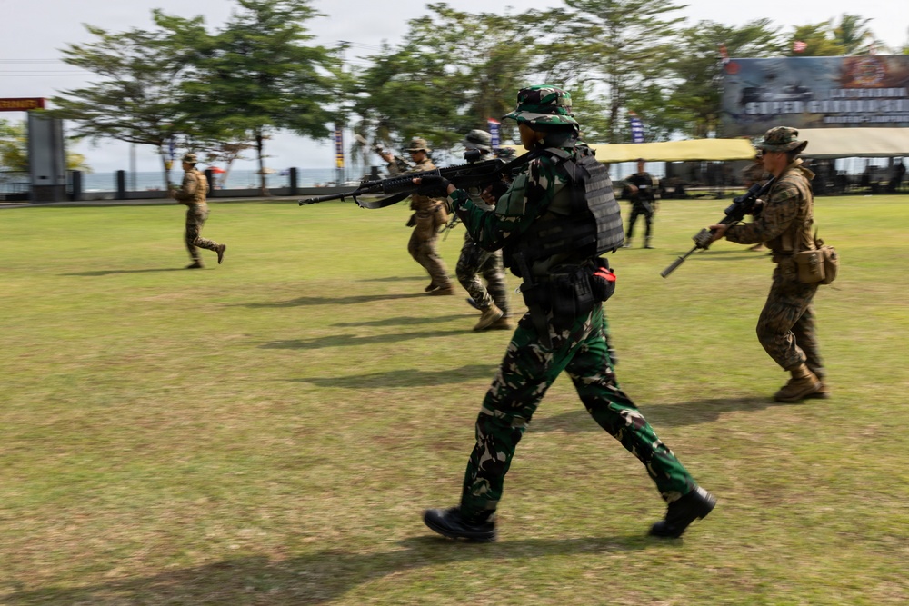 The image shows military personnel in mixed uniforms conducting what appears to be a joint training exercise on an open grass field. Soldiers wearing Indonesian TNI (Tentara Nasional Indonesia) camouflage and others in tan/MultiCam uniforms are moving tactically with rifles. The motion blur in the image suggests dynamic movement during the exercise.