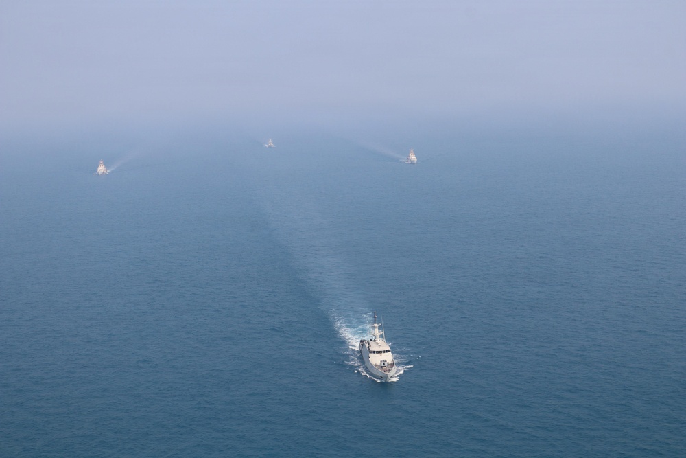 An aerial photograph shows a formation of four vessels navigating open sea, with one larger white vessel in the foreground leading and three smaller vessels arranged in a V-formation behind it. The three rear vessels appear to be spraying water arcs, suggesting a ceremonial or escort formation. The image is taken from a significant altitude with hazy atmospheric conditions reducing visibility in the background.