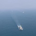 An aerial photograph shows a formation of four vessels navigating open sea, with one larger white vessel in the foreground leading and three smaller vessels arranged in a V-formation behind it. The three rear vessels appear to be spraying water arcs, suggesting a ceremonial or escort formation. The image is taken from a significant altitude with hazy atmospheric conditions reducing visibility in the background.