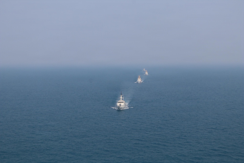 An aerial or elevated view of multiple naval vessels sailing in a loose column formation across open ocean. The lead vessel is a white-hulled ship, likely a coast guard or patrol vessel, followed by two or three darker warships in the distance. The scene appears to depict a naval exercise or coordinated patrol operation.