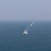 An aerial or elevated view of multiple naval vessels sailing in a loose column formation across open ocean. The lead vessel is a white-hulled ship, likely a coast guard or patrol vessel, followed by two or three darker warships in the distance. The scene appears to depict a naval exercise or coordinated patrol operation.
