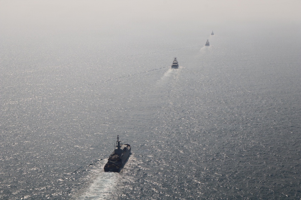 An aerial photograph shows a formation of five naval vessels sailing in a single-file line across open, calm sea. The ships are evenly spaced and appear to be traveling in the same direction, leaving visible wake trails behind them. Hazy atmospheric conditions reduce visibility of the more distant vessels.