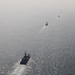 An aerial photograph shows a formation of five naval vessels sailing in a single-file line across open, calm sea. The ships are evenly spaced and appear to be traveling in the same direction, leaving visible wake trails behind them. Hazy atmospheric conditions reduce visibility of the more distant vessels.