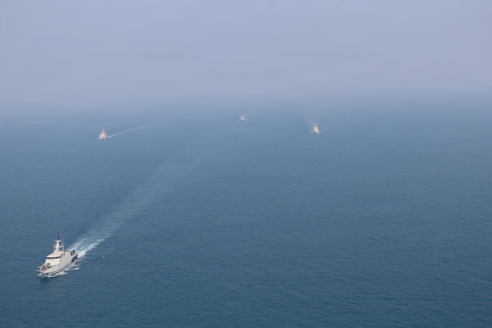 An aerial photograph shows a formation of five naval or coast guard vessels navigating across open blue-gray water in hazy conditions. The lead vessel is clearly visible in the foreground with a white wake trailing behind it, while four additional vessels are arranged in a diagonal formation extending into the misty background. The image appears to depict a coordinated maritime patrol or exercise.