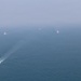An aerial photograph shows a formation of five naval or coast guard vessels navigating across open blue-gray water in hazy conditions. The lead vessel is clearly visible in the foreground with a white wake trailing behind it, while four additional vessels are arranged in a diagonal formation extending into the misty background. The image appears to depict a coordinated maritime patrol or exercise.