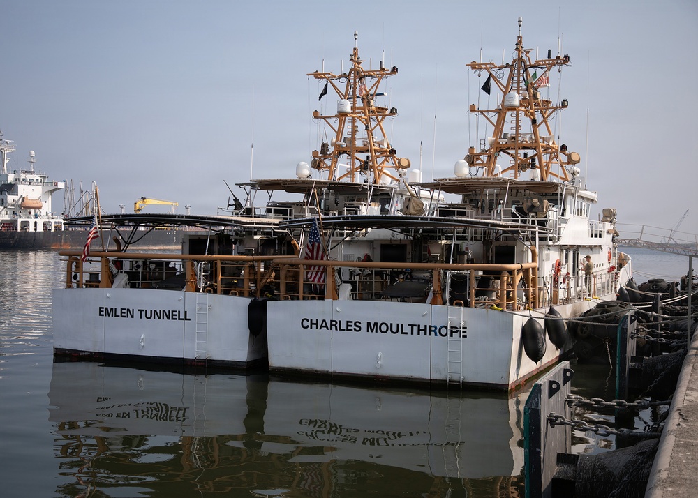 Two white vessels named 'Emlen Tunnell' and 'Charles Moulthrope' are docked side by side at a pier. Both ships appear to be similar in design, featuring prominent tan/orange-colored mast structures with navigation and communication equipment. American flags are visible on both vessels, and their names are reflected in the calm harbor water below.
