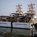 Two white vessels named 'Emlen Tunnell' and 'Charles Moulthrope' are docked side by side at a pier. Both ships appear to be similar in design, featuring prominent tan/orange-colored mast structures with navigation and communication equipment. American flags are visible on both vessels, and their names are reflected in the calm harbor water below.
