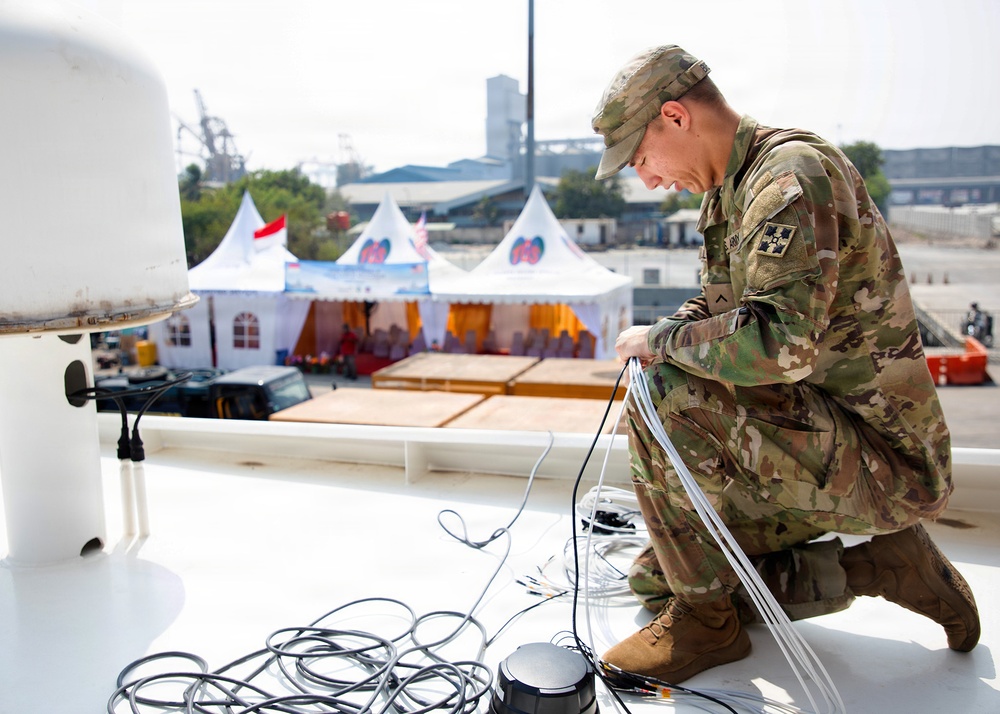 A U.S. Army soldier in OCP (Operational Camouflage Pattern) uniform is kneeling on what appears to be the deck of a ship or elevated platform, working with cables and wiring equipment. In the background, a ceremonial or event area is visible with white peaked tents decorated with what appears to be Indonesian and American flags, suggesting a bilateral military or diplomatic event. The setting appears to be a port or harbor facility.