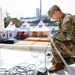A U.S. Army soldier in OCP (Operational Camouflage Pattern) uniform is kneeling on what appears to be the deck of a ship or elevated platform, working with cables and wiring equipment. In the background, a ceremonial or event area is visible with white peaked tents decorated with what appears to be Indonesian and American flags, suggesting a bilateral military or diplomatic event. The setting appears to be a port or harbor facility.
