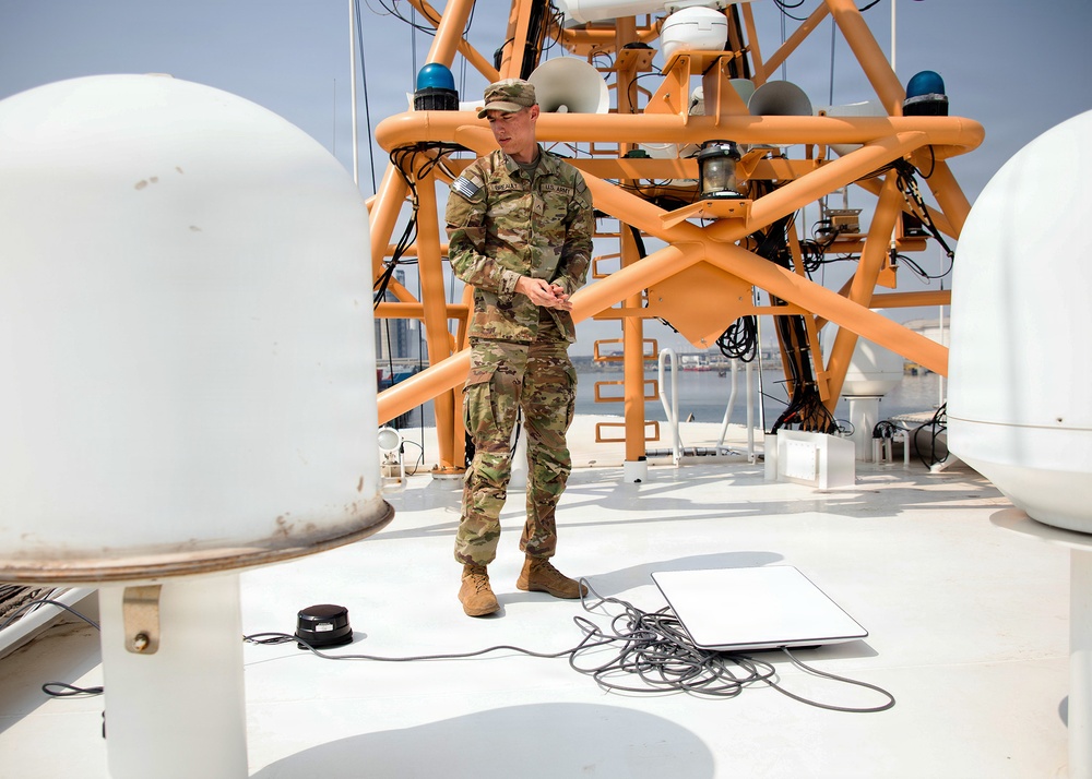 A U.S. Army soldier in OCP (Operational Camouflage Pattern) uniform stands on the upper deck of a vessel, surrounded by communication and navigation equipment. The soldier appears to be conducting equipment checks or a technical assessment near cables, a laptop, and satellite dome antennas. The setting appears to be a port or harbor environment based on the visible water and infrastructure in the background.