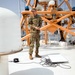 A U.S. Army soldier in OCP (Operational Camouflage Pattern) uniform stands on the upper deck of a vessel, surrounded by communication and navigation equipment. The soldier appears to be conducting equipment checks or a technical assessment near cables, a laptop, and satellite dome antennas. The setting appears to be a port or harbor environment based on the visible water and infrastructure in the background.