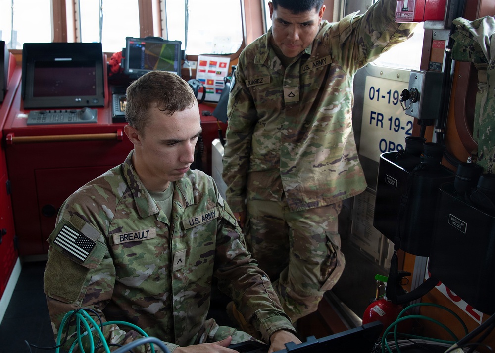 Two U.S. Army soldiers are working together at what appears to be a ship's bridge or command station. The soldier in the foreground, identified by name tape as 'BREAULT,' is operating a laptop or console, while the soldier standing behind him, identified as 'JUAREZ,' points toward equipment on the right side. Both wear OCP (Operational Camouflage Pattern) uniforms with U.S. Army insignia and American flag patches.