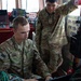 Two U.S. Army soldiers are working together at what appears to be a ship's bridge or command station. The soldier in the foreground, identified by name tape as 'BREAULT,' is operating a laptop or console, while the soldier standing behind him, identified as 'JUAREZ,' points toward equipment on the right side. Both wear OCP (Operational Camouflage Pattern) uniforms with U.S. Army insignia and American flag patches.