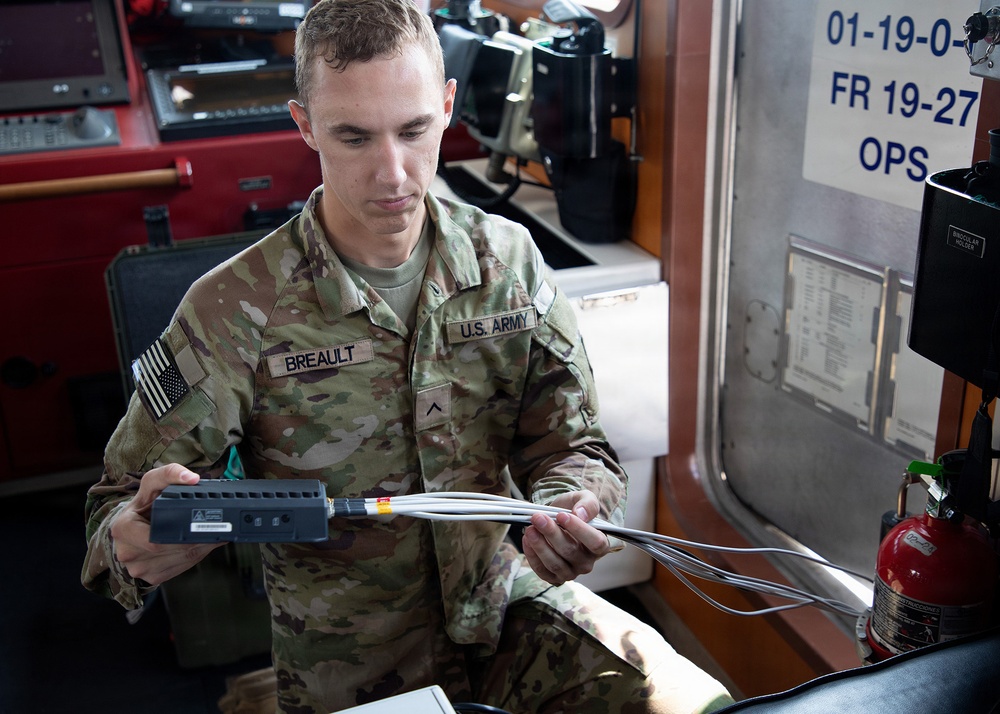 A U.S. Army soldier named Breault, wearing Operational Camouflage Pattern (OCP) uniform, is handling a small electronic device connected to multiple cables inside what appears to be a military operations vehicle or command post. He appears to be inspecting or connecting communications or electronic equipment. The setting suggests a field operations or training environment.